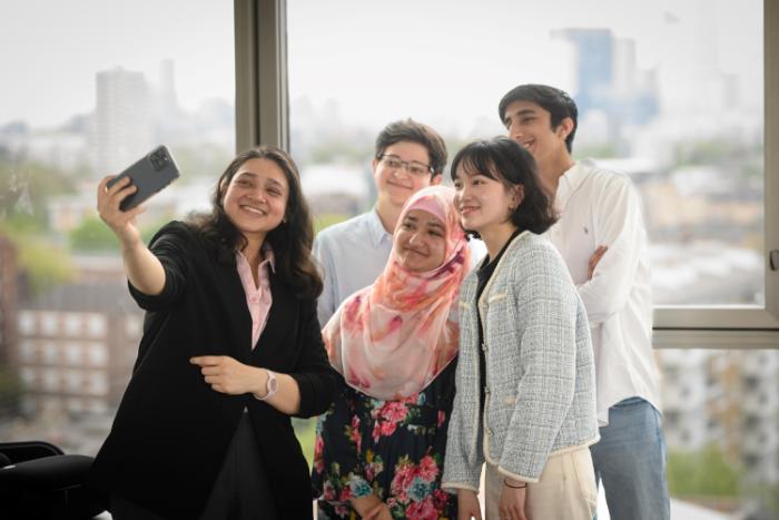 A group of five interns by a window, one of them is taking a selfie of the group