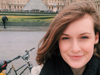 Eliza smiling at the camera in Paris, she is in front of the Louvre museum