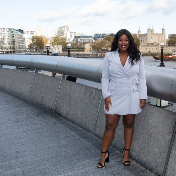 Photo of alumna, Paula Olugbemi standing by the River Thames in London. she is wearing a white dress with black heels and is smiling at the camera.