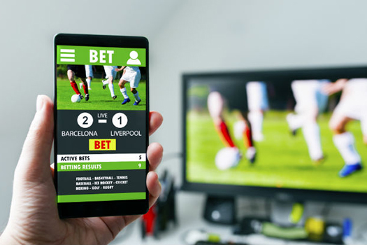A hand holding up a mobile phone displaying a bet they've made on a football (soccer) game A TV is out of focus in the background playing the game