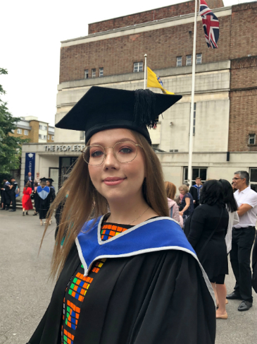 Karina stands outdoors wearing a graduation cap and gown and underneath that a brightly patterned dress with orange and green on it. She wears delicate gold reading glasses. Her dark blonde hair is loose and blows out behind her.
