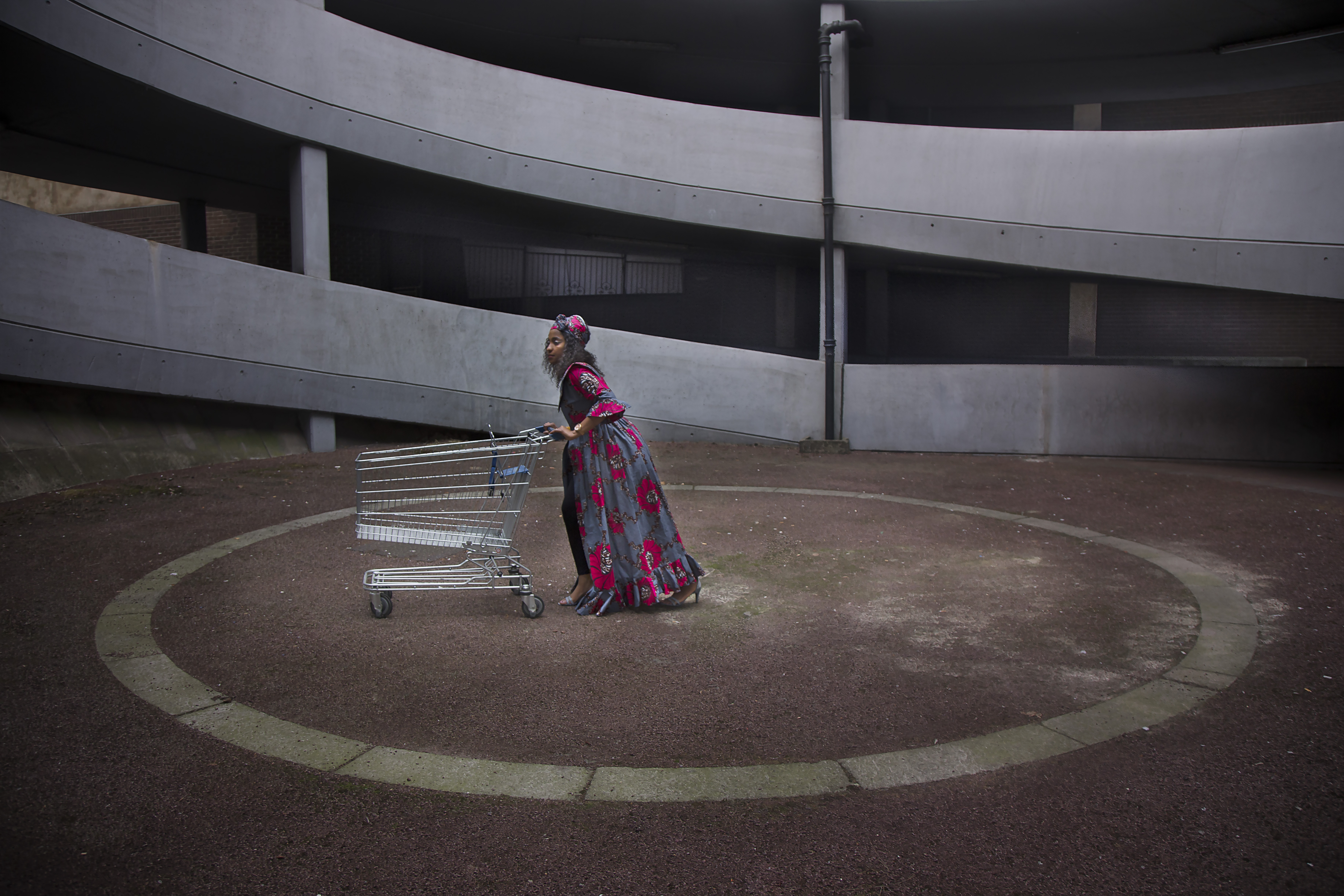 A women in a long pink and grey coat and strappy silver sandals pushes a shopping trolley across the centre of a spiraling concrete ramp