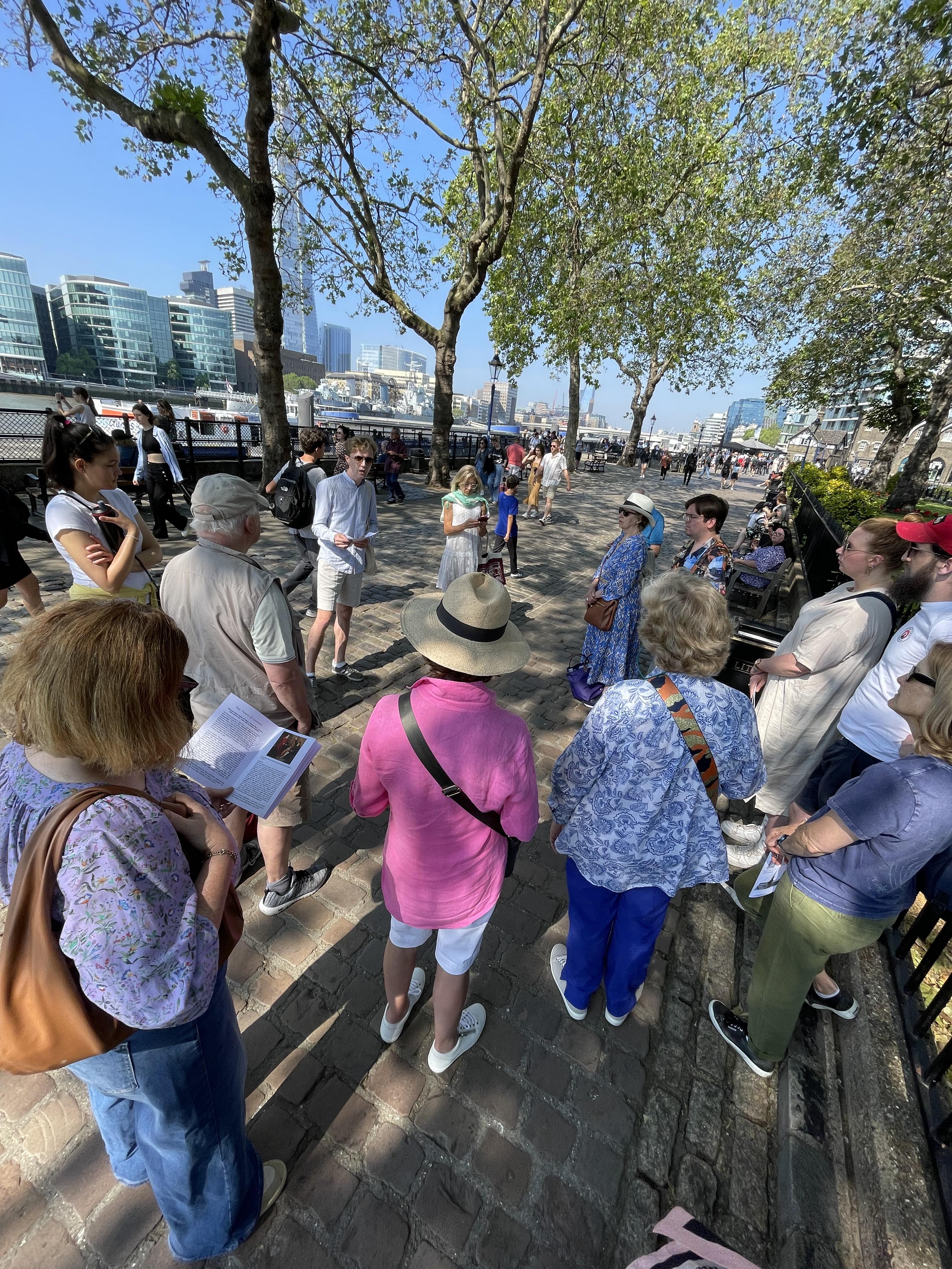Alexander Gould, a white man with curly brown hair, presenting to the tour group on the bank of the Thames next to the Tower of London. In the background is the river, buildings including the Shard, large trees and blue sky.