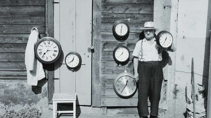 Black and white photo of a man standing in front of a number of analogue clocks each showing different times