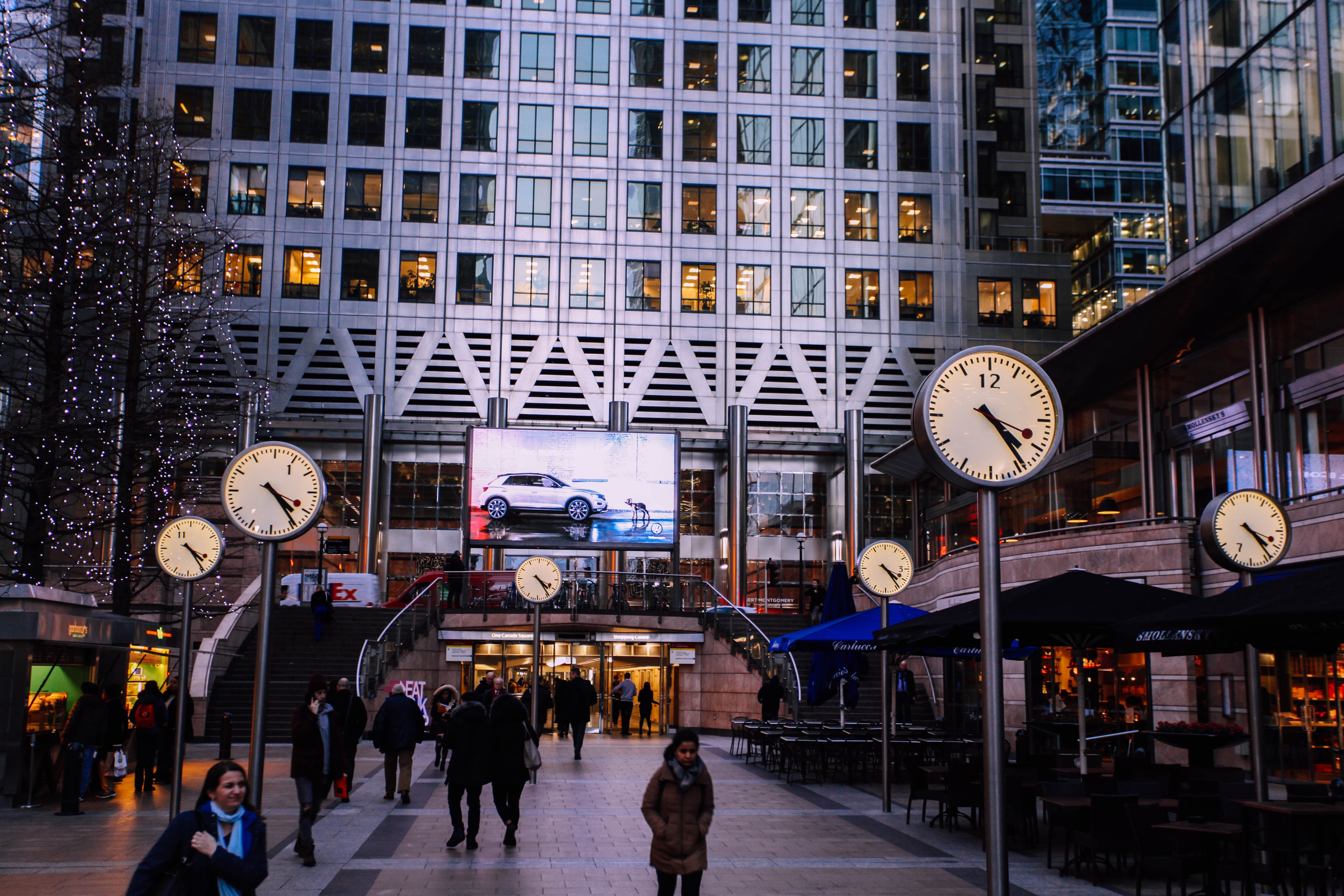 A view of Canary Wharf showing people, buildings and large illuminated clockfaces