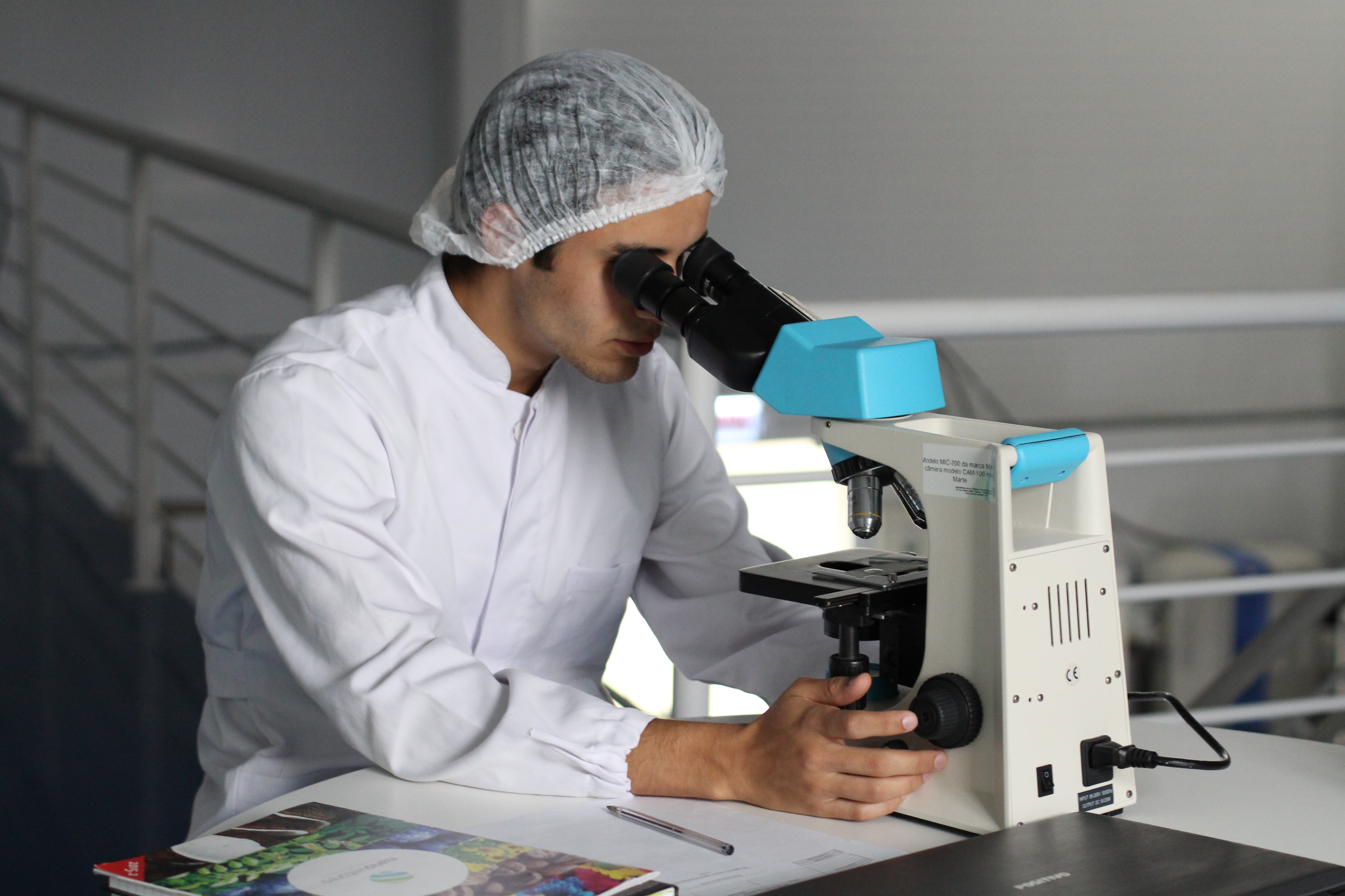 man in white lab coat and hair net sitting down and looking in microscope.