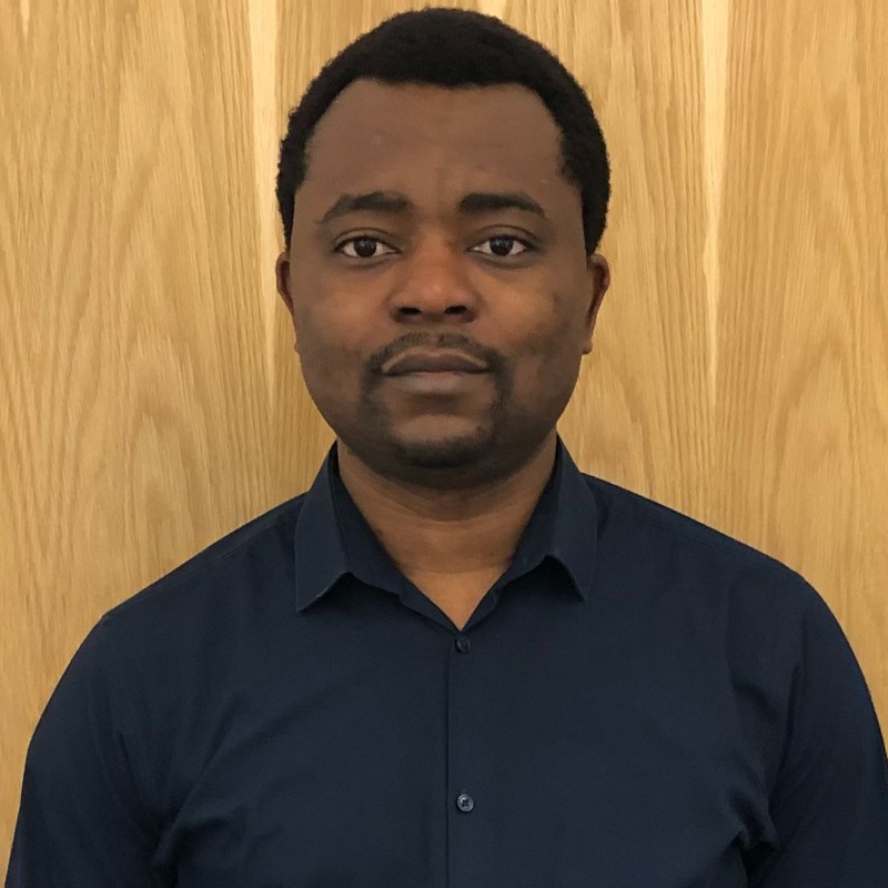 Headshot of Solomon Odafe Akpotozor. He is wearing a black shirt and standing in front of a light wooden background.