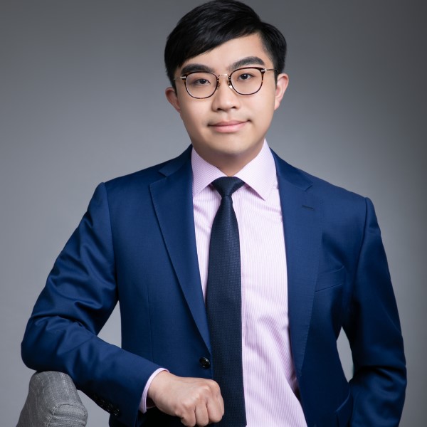 Headshot of alumnus, Kelvin Mak, wearing glasses, a smart navy blue suit, with a pale pink shirt and navy tie. His elbow is propped on a chair and he is smiling at the camera.