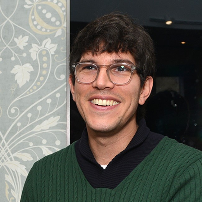 Headshot of alumnus, Mark Bergfeld. He is wearing a green and black jumper and glasses and is smiling at someone off camera.