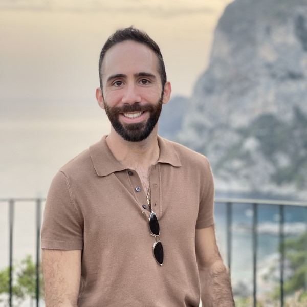 Photo of Peris Anastasiou standing in front of a view of sea and mountains. He is wearing a beige t-shirt and is smiling at the camera.