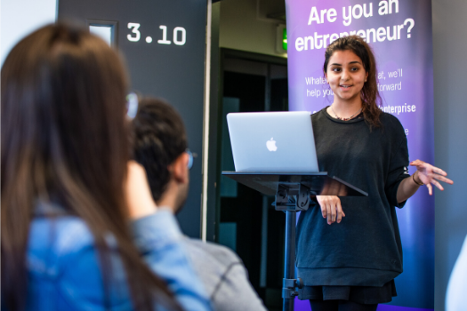 A female student  standing speaking to an audience with a computer screen in front of her 