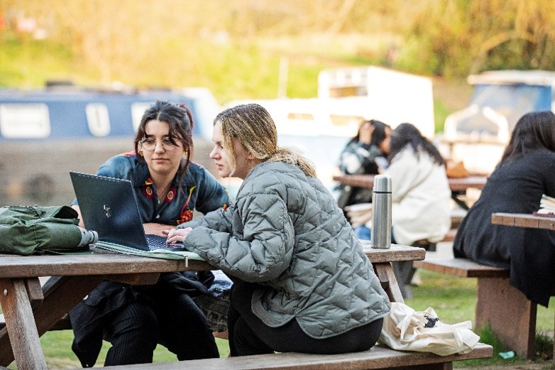 Two students at a picnic table looking at a laptop, canal boats are visible behind them