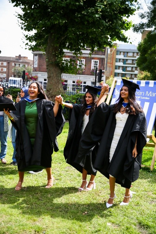 Three students in graduation robes and mortarboards holding hands and jumping joyfully
