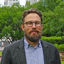 Man with beard and glasses in casual blue shirt and tweed jacket smiling