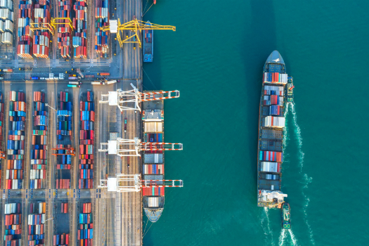 An image from above of a cargo ship sailing on the sea adjacent to docks loaded with shipping containers.