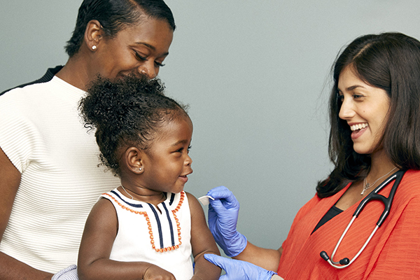 A toddler sits on her mum's knee, smiling at the doctor. The doctor is wearing gloves and is cleaning the toddler's arm ready for an injection.