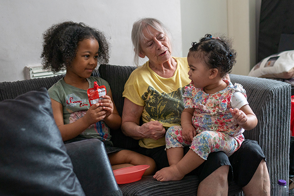 A woman sits with her two young grandchildren on the sofa, with the little one on her knee