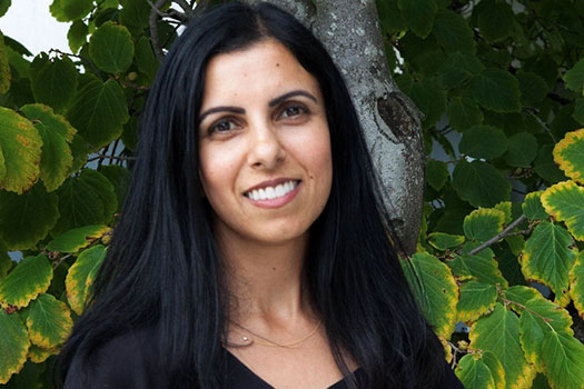 An image of Professor Renisa Mawani. She stands outdoors with a tree and foliage behind her. She has light brown skin, brown eyes, long straight black hair and wears a black v-neck top. She is smiling.
