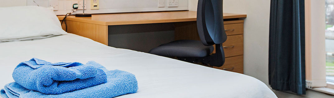 The slider shows the layout of one of our student bedrooms, blue towels on the bed, a desk, a desk chair and part of the window. It has the heading: summer accommodation at QMUL