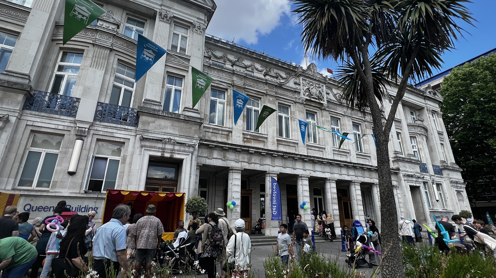 A classical style building in the background. People are festively gathered in the square in front of it. The square is decorated with green and blue bunting which reads: Festival of Communities.