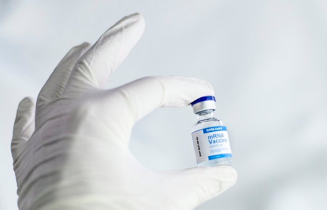 A hand wearing a white medical glove holding a small vial of mRNA Covid-19 vaccine