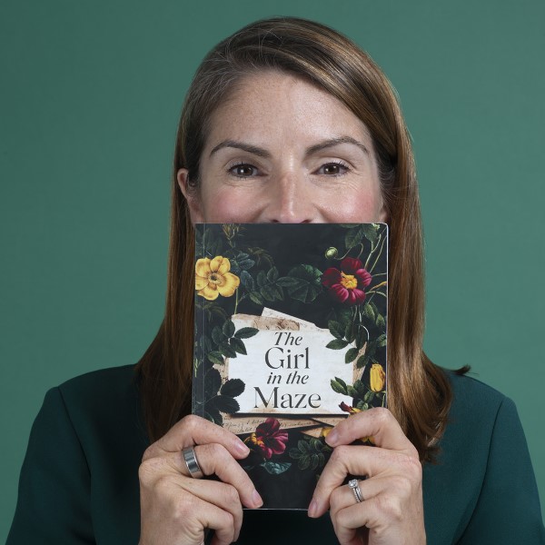 Photo of alumna, Cathy Hayward, holding her debut novel, The Girl in the Maze, with two hands. The book is obscuring the bottom half of her face and she is standing in front of a dark green background which compliments the book cover.