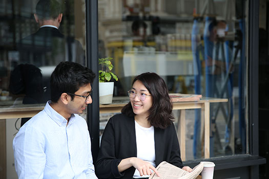 One male student and one female student chatting outside a coffee shop. The woman is wearing glasses, and a black jacket and holding a piece of paper. He is wearing glasses and a white shirt.