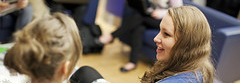 Two women sit together having a conversation. The woman on the left has her back to the camera. The woman on the right has long dark blonde hair and we see her face in profile. She is wearing a blue cardigan.