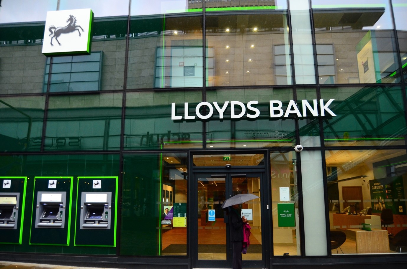 The front of a Lloyds bank branch. A person stands outside the doors under an umbrella.
