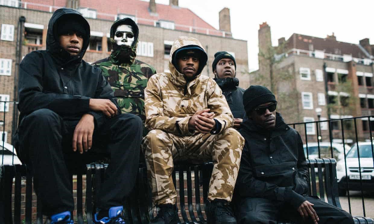 Four young black men sit together on a bench. They are dressed casually wearing tracksuits, hoodies and wool hats. One of the men wears sunglasses. One man stands at the back wearing a silver mask.