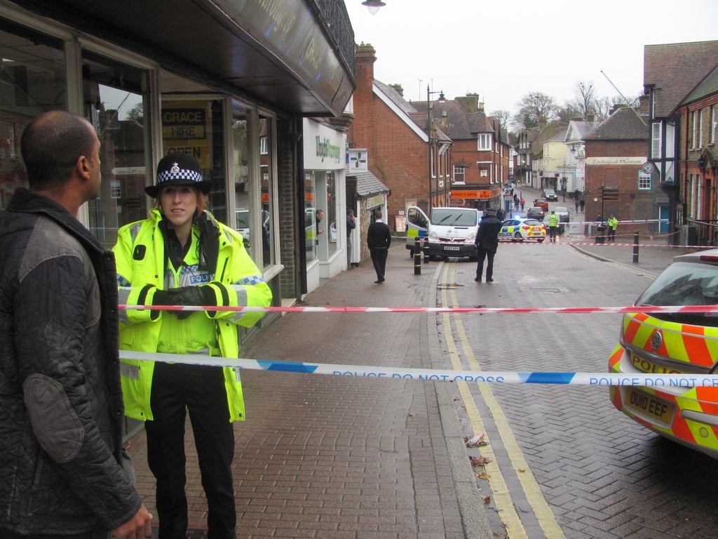 A female police officer stands at a police cordon set up on a street. There is a man standing on the other side of the cordon opposite her with his back to the camera. In the distance there is a police van and beyond that a police car.