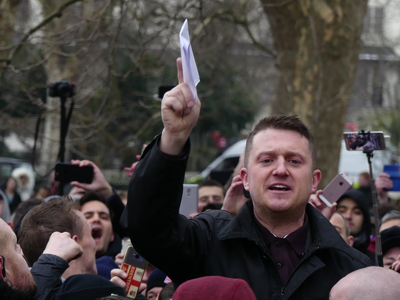 A picture of right-wing activist and politician Tommy Robinsonstanding amongst a crowd of people, many of whom are using their phones to film/take pictures of him