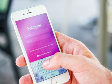 A close-up image of a hand holding a white Apple mobile phone. The Instagram login screen is displayed on the phone.