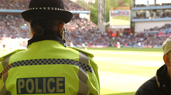 Police officer standing with their back to the camera in a hi-viz jacket with the word police on the back. There is a football pitch and crowd in the background