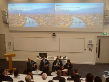 A shot taken from the back of a lecture theatre looking down on a panel of four people - three men and one women. On the screens in front of the audience there is a picture of the London landscape and the Thames with the text 