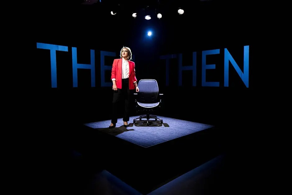 A woman in a red blazer standing on a small stage next to an office chair.