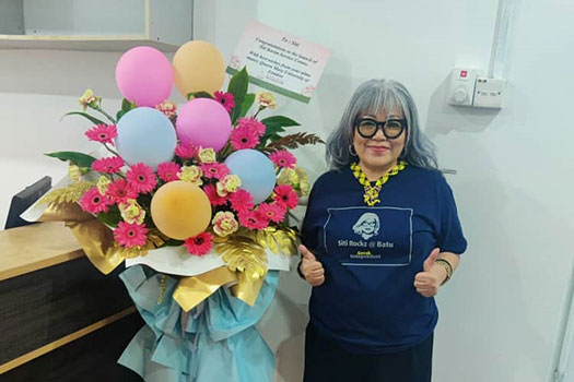 Siti Kasim in a blue dress, black rimmed glasses, standing next to a bouquet of flowers