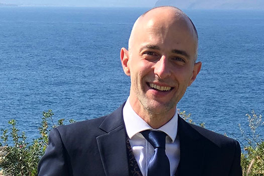 Ioannis Kokkoris wearing a suit and blue tie, standing in front of the ocean