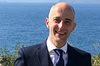 Ioannis Kokkoris wearing a suit and blue tie, standing in front of the ocean