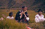 A man sat on the grass playing a wind instrument with a woman and child. They are wearing traditional dress.