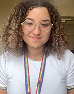 Head and shoulders photo of Malak Benslama Dabdoub. She has medium length curly hair, and is wearing clear glasses, a white t-shirt and a rainbow lanyard.