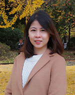 Puangrat Patomsirirak standing in a park wearing a tan coat over a white jumper