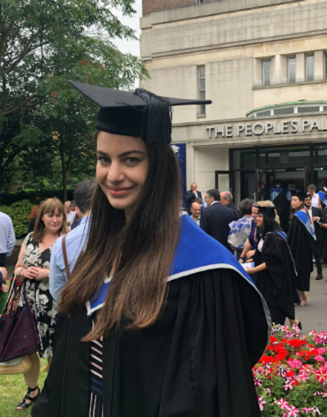 A picture of Sylvia in graduation gown and cap at the School of Law's graduation ceremony in July 2019. She has long medium straight brown hair and fair skin. She is smiling. The People's Palace can be seen in the background.
