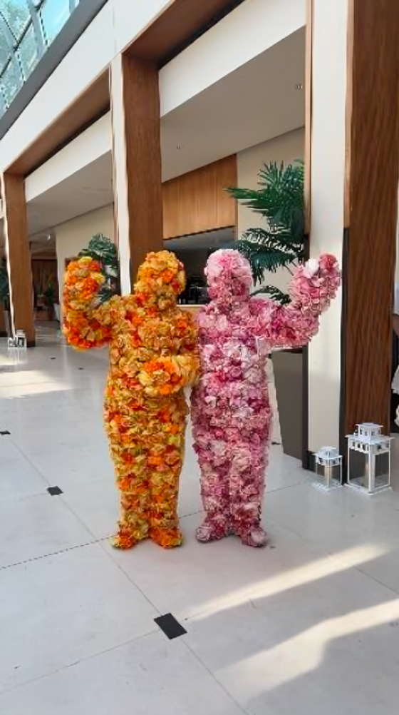 Two people covered from head to toe in flowers. The other person is covered in orange flowers and the other one in pink