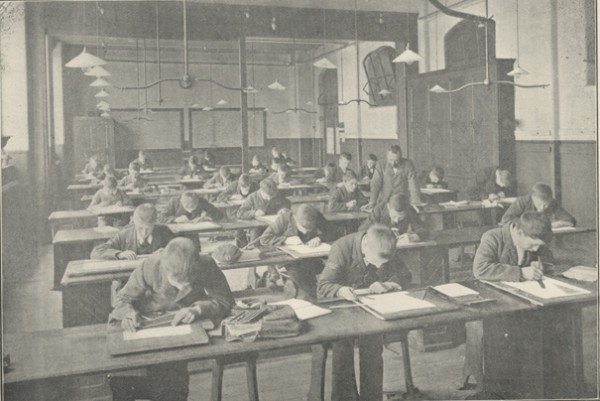 Black and white photograph from about 1900 of a classroom in the technical schools. Young boys sit at desks with their heads down drawing and using rulers. 