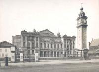 Black and white photograph of the front of the People's Palace in Mile End in the early twentieth century showing a cobbled street, clock tower, a large stone building, the winter garden to the right, st benets church to the left and posters for events on a notice board
