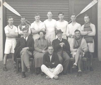 Black and white photograph of the Queen Mary Rowing Eight from 1938.  The group is arranged in three rows, one man in the front is seated on the ground, one woman and three men are seated on chairs behind him, and seven men stand in a row at the back