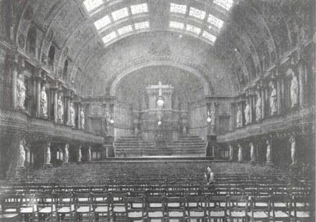 Black and white photograph of the Queen's Hall in the 1890s.  There is a stage, with rows of seats in the foreground. On each side of the hall there are columns of statues of different women from history