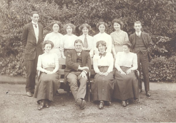 Black and white photograph from the late 1800s depicting a group of men and women in two rows.  Four people sit in the front row with seven people standing behind.