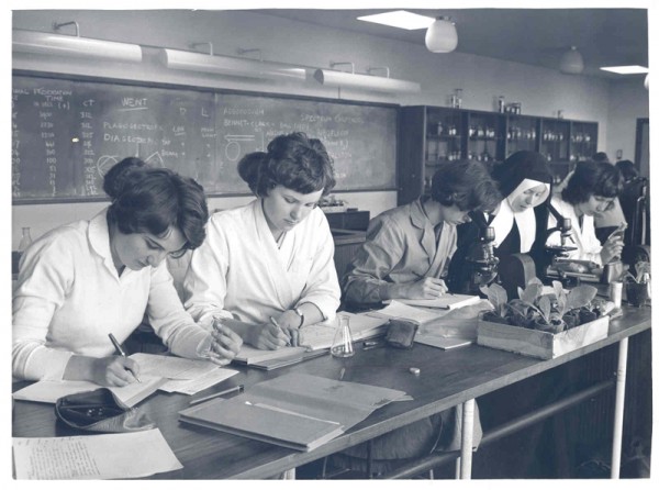 Women students in 1962 study in a laboratory.  A nun in black habit can be seen in the middle of the picture and there are microscopes on the benches